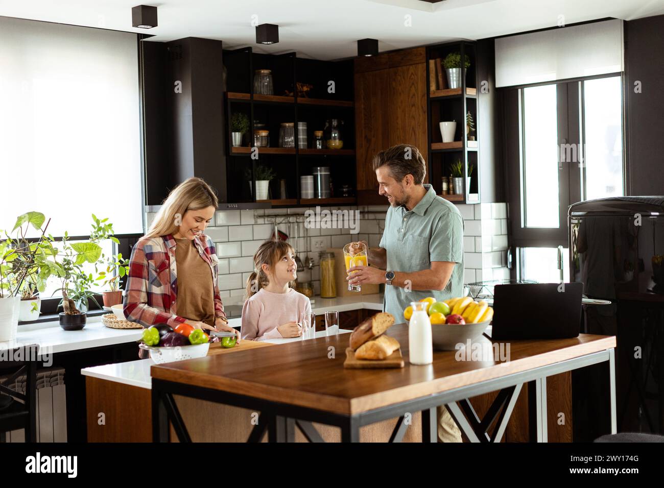 Young family chatting and preparing food around a bustling kitchen counter filled with fresh ingredients and cooking utensils Stock Photo