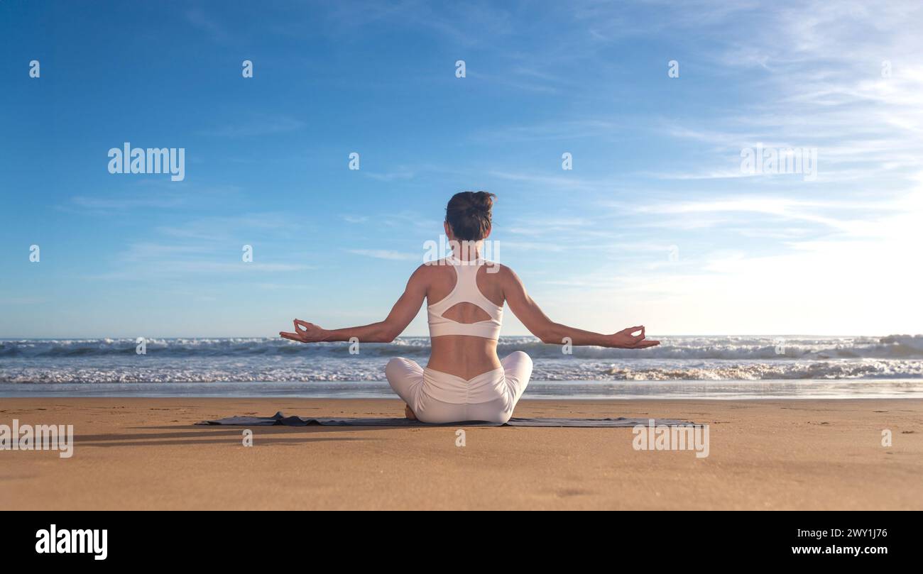 Outdoor yoga. Woman meditating, sitting in lotus position on fitness mat on beach, practicing meditation near ocean, back view Stock Photo