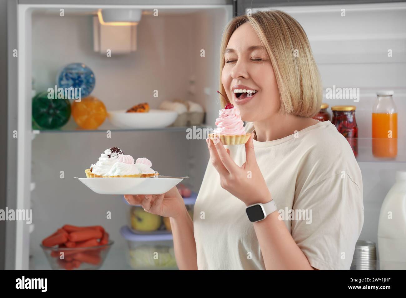 Beautiful woman with tasty cakes near fridge in kitchen. Overeating concept Stock Photo