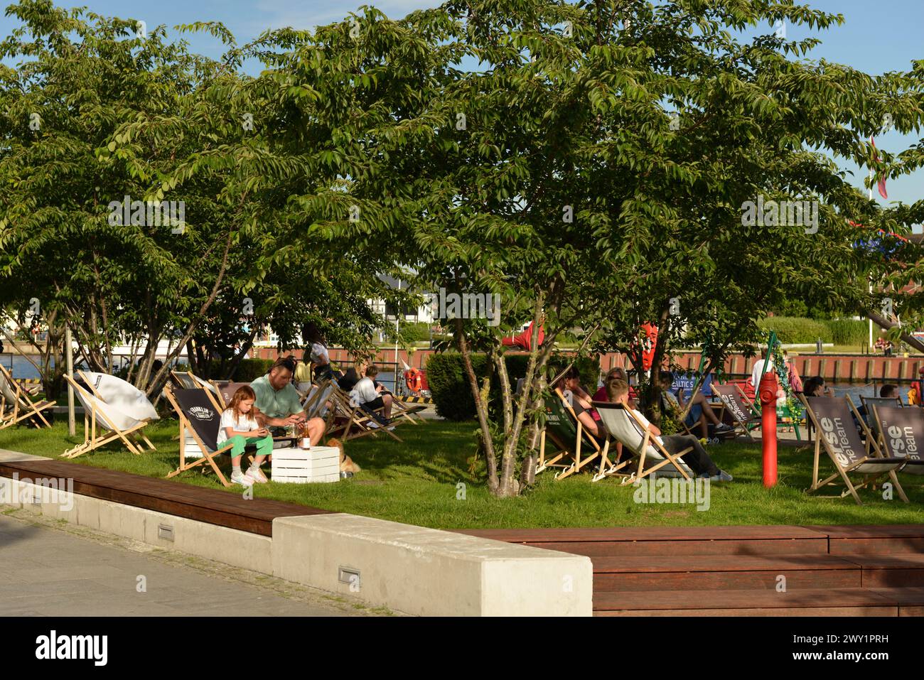 Tourists dining alfresco in deckchairs under cherry trees at a residential complex by Invest Komfort at Motlawa River in the Old Town of Gdansk, Polan Stock Photo