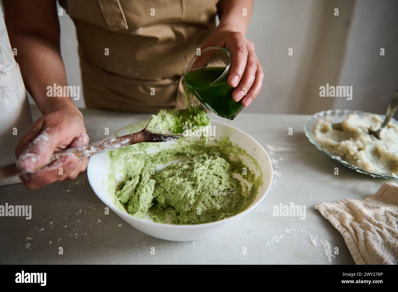 Close-up hands of a chef pastry, housewife pouring green spinach water into a bowl with wheat four, making dough of green color for ravioli or dumplin Stock Photo