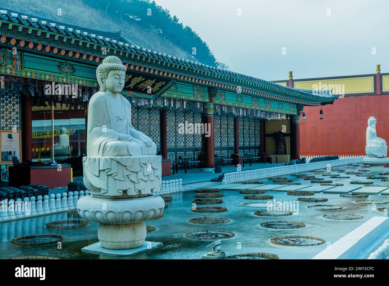 Large stone carved seated Buddha in frozen man made pond with temple building in background in Yeongcheon, South Korea Stock Photo