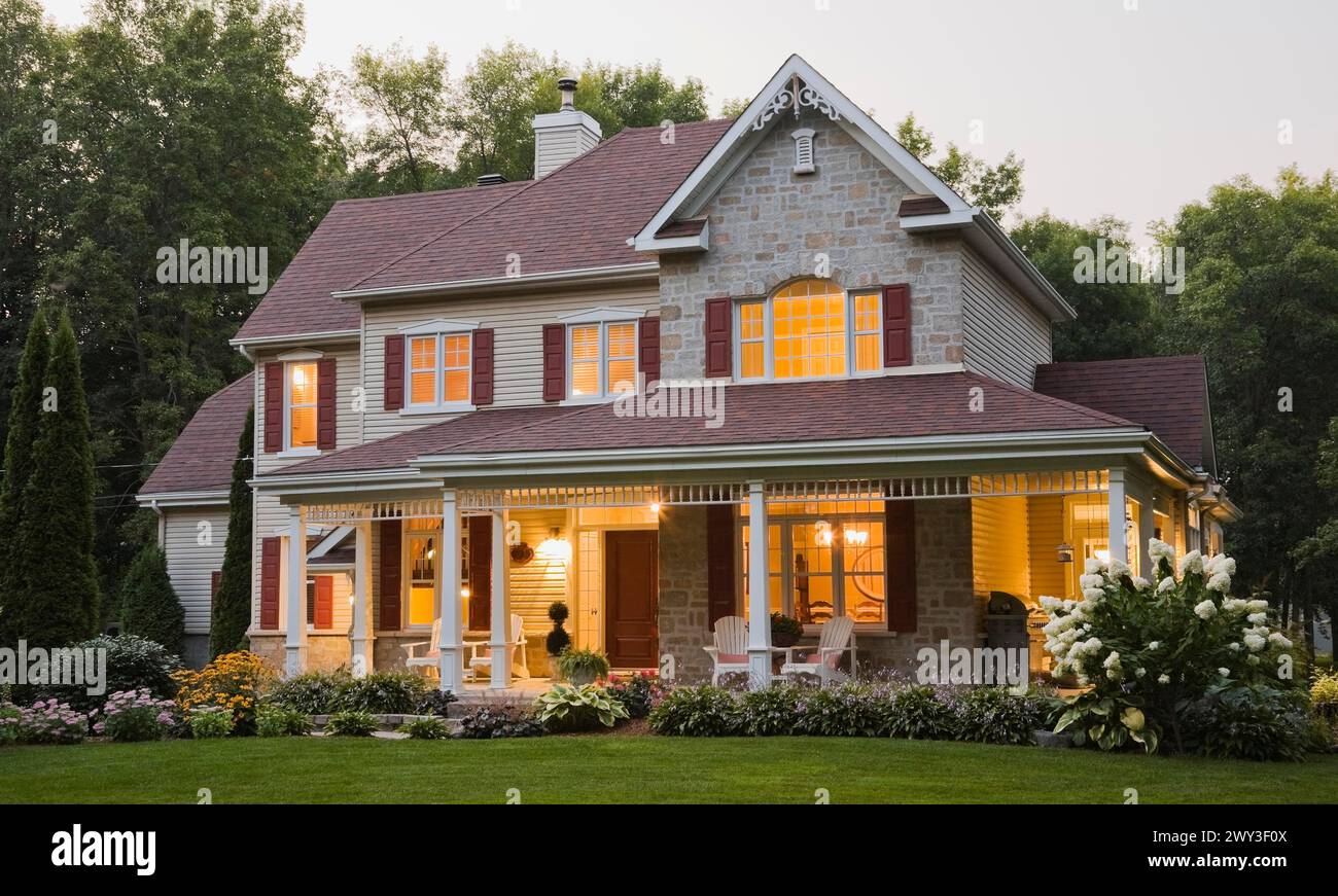 Large illuminated two story beige and tan cut stone home exterior with white and red trim, multiple burgundy asphalt shingles roofs plus Adirondack Stock Photo