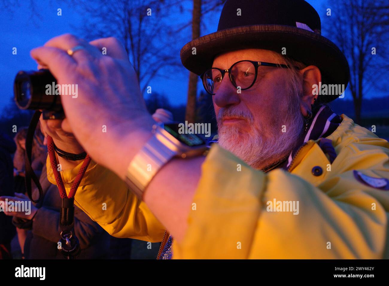 Older male vlogger in hat films the Easter bonfire. His face is lit red by the fire Stock Photo