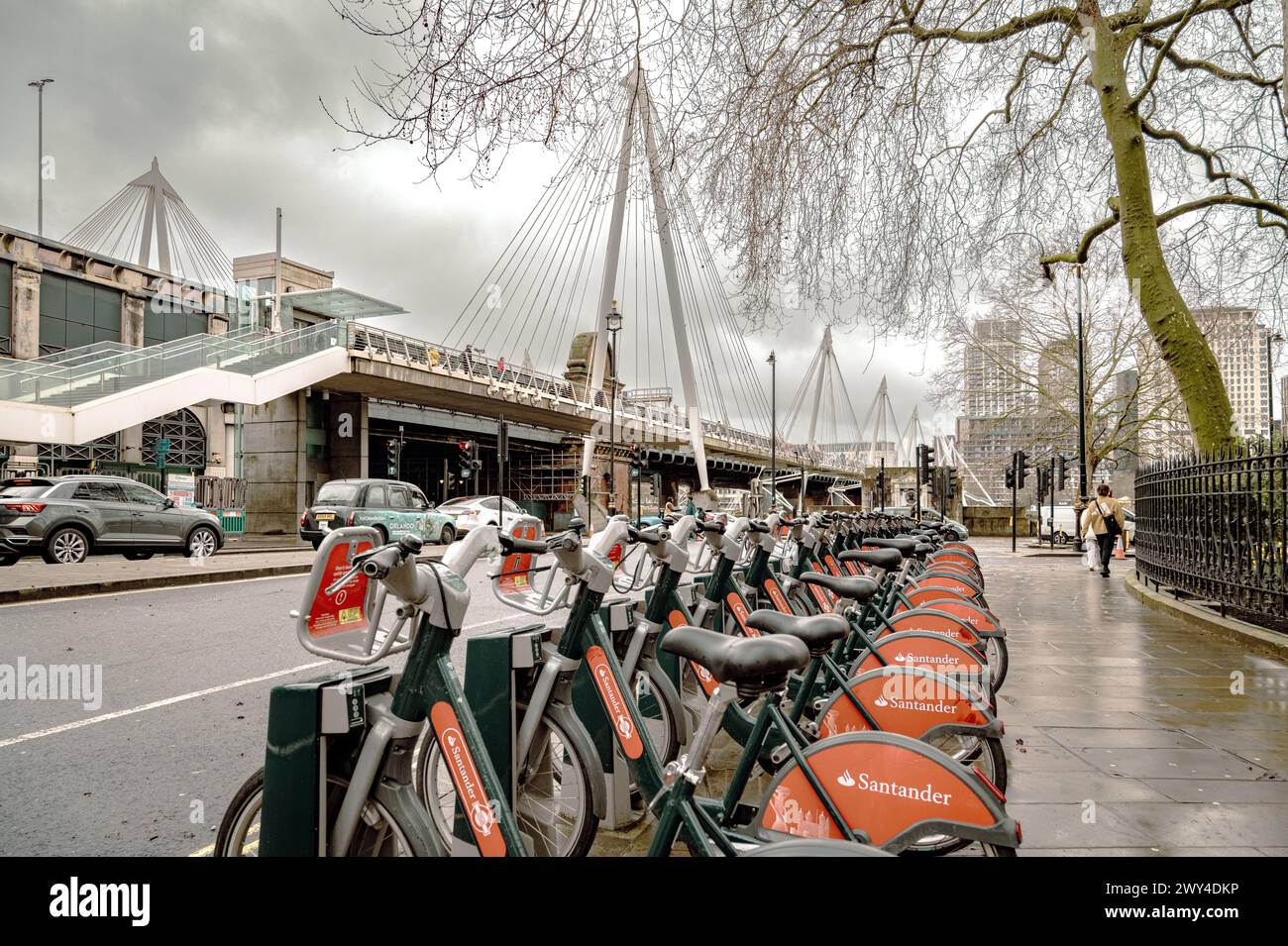 A row of Santander E-Bikes for hire in Central London. In the background pedestrians are crossing the River Thames on the Golden Jubilee Bridge. Stock Photo