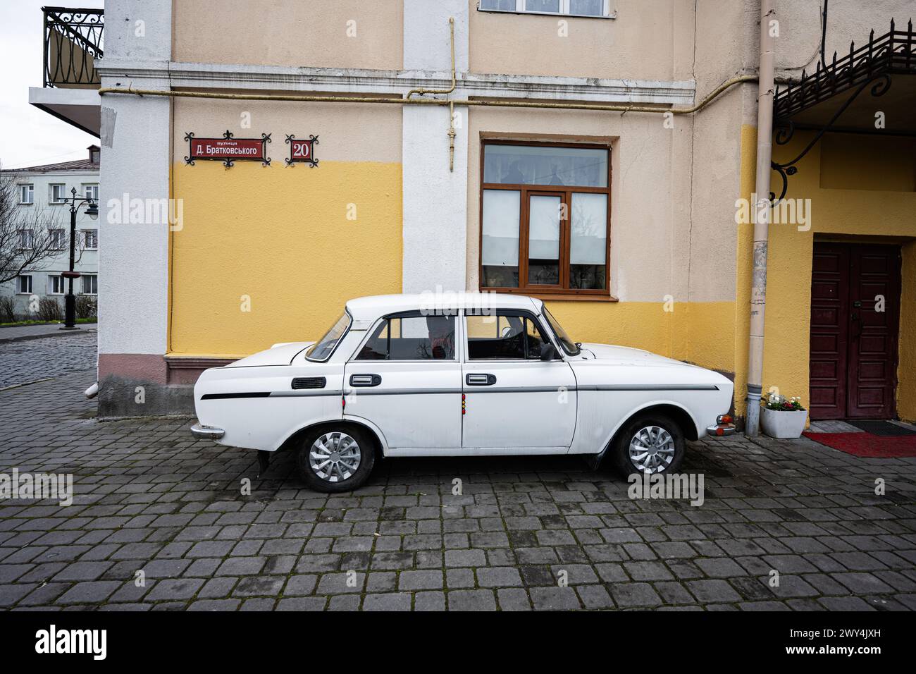 Lutsk, Ukraine - March, 2024: A classic white soviet Moskvitch sedan car carefully parked by a colorful building facade with traditional European arch Stock Photo