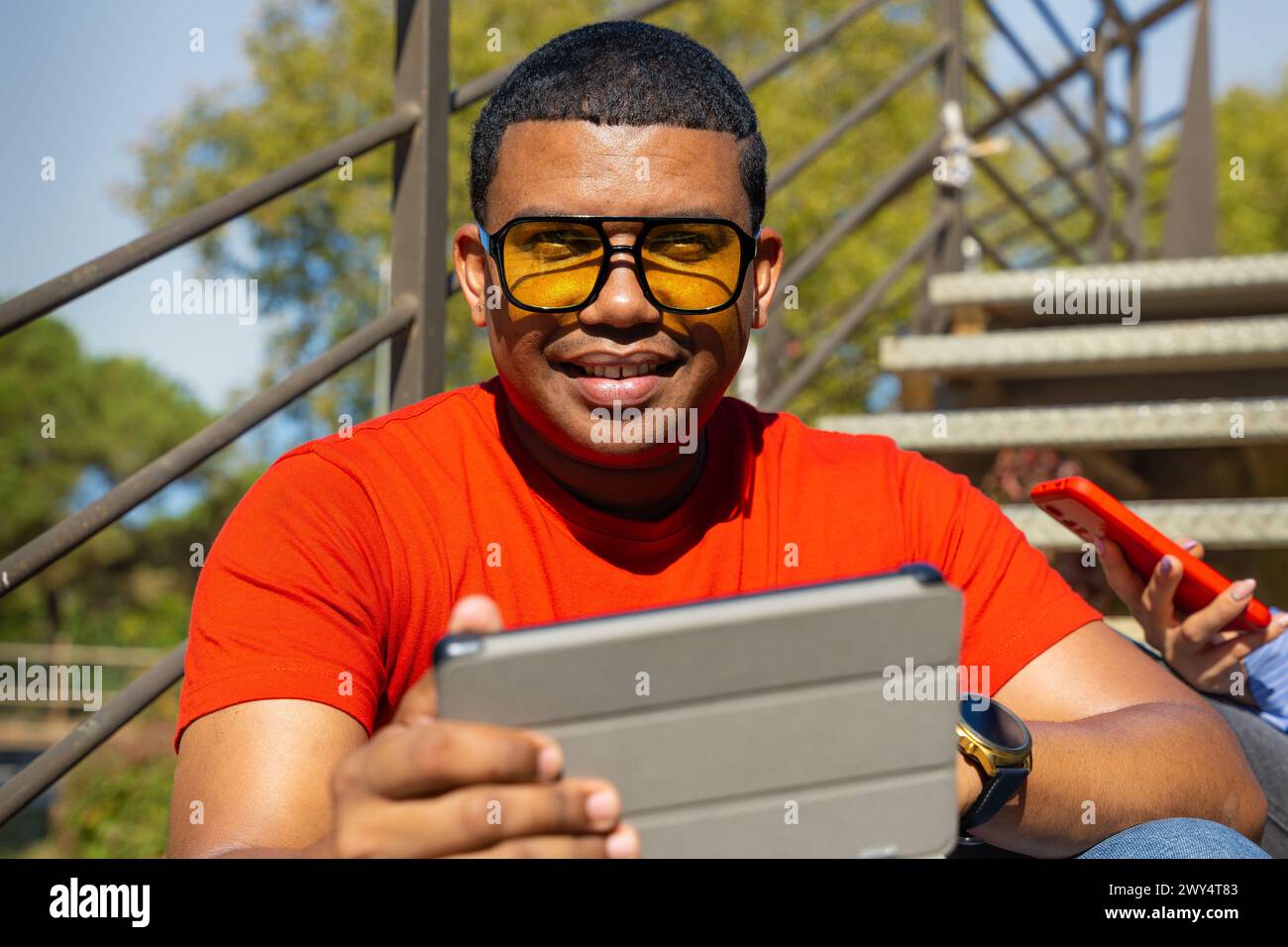 Glamorous smiling young man, sitting using a app on a digital tablet outdoors. Stock Photo