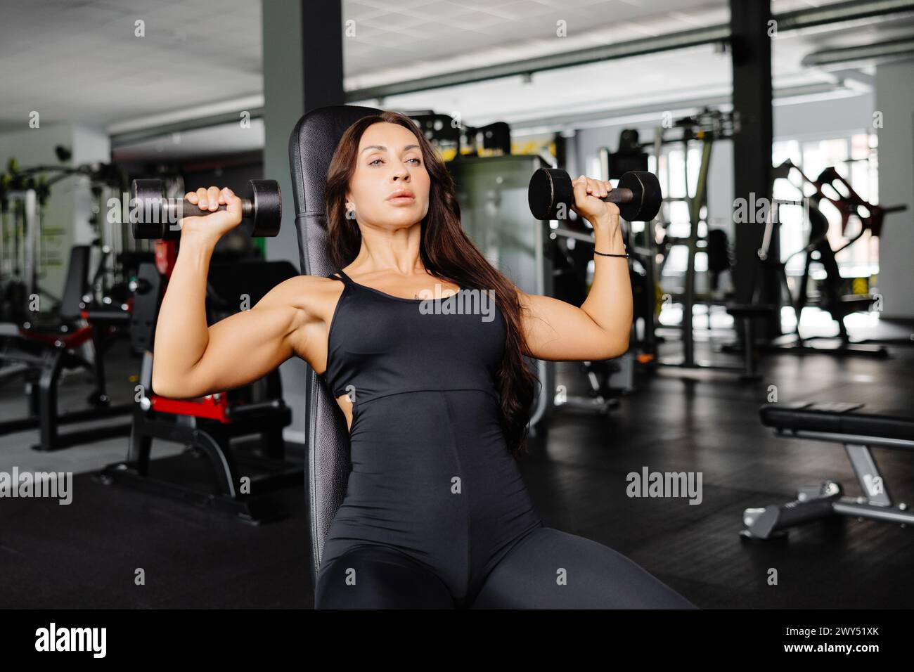 Young fit woman practicing dumbbell press while sitting on a bench. Slim female in sportswear exercises with weights in a gym. Stock Photo