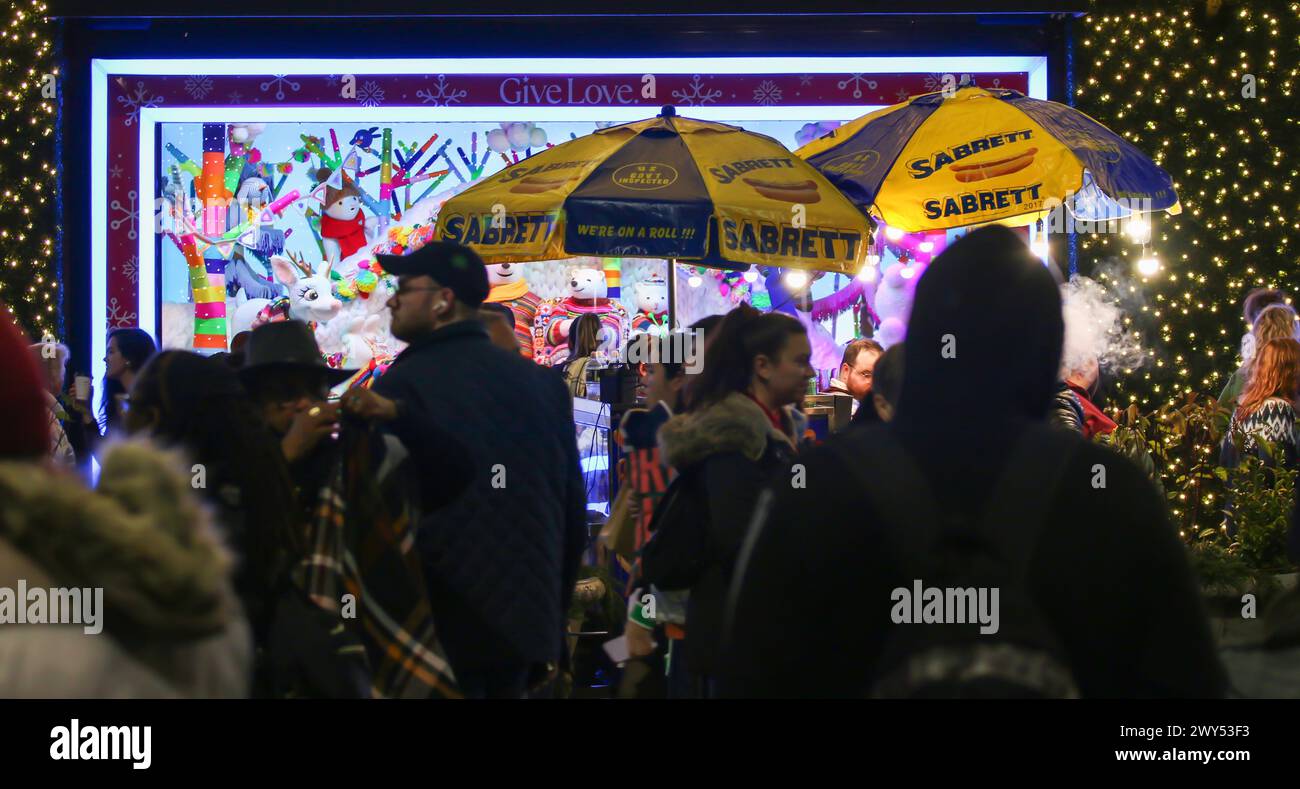 New York City, New York, USA - 3 December 2023: A group of individuals standing on sidewalk with hot dog vendors umbrellas outside a store. Stock Photo