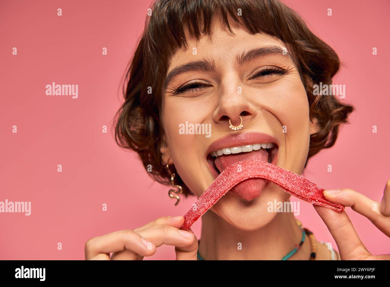 happy and young woman with nose piercing licking sweet and sour candy strip on pink background Stock Photo