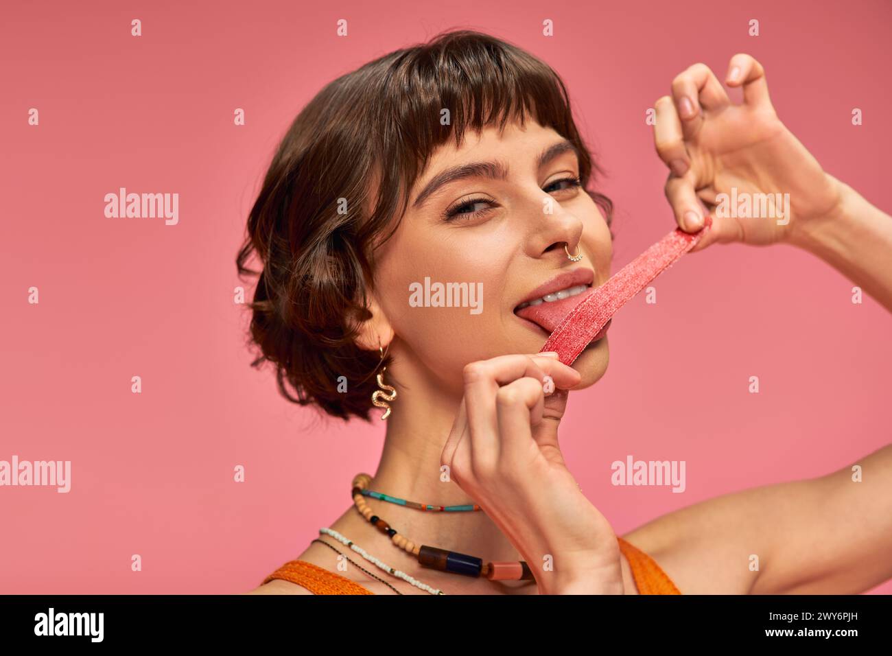 cheerful and young woman with nose piercing licking sweet and sour candy strip on pink background Stock Photo
