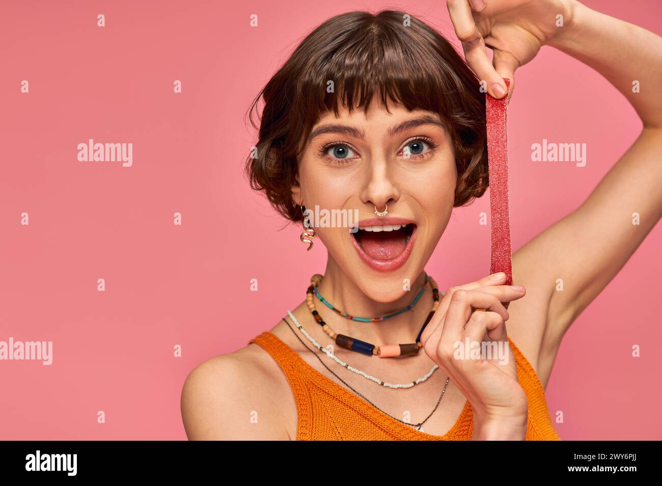 excited and young woman with nose piercing holding sweet and sour candy strip on pink background Stock Photo