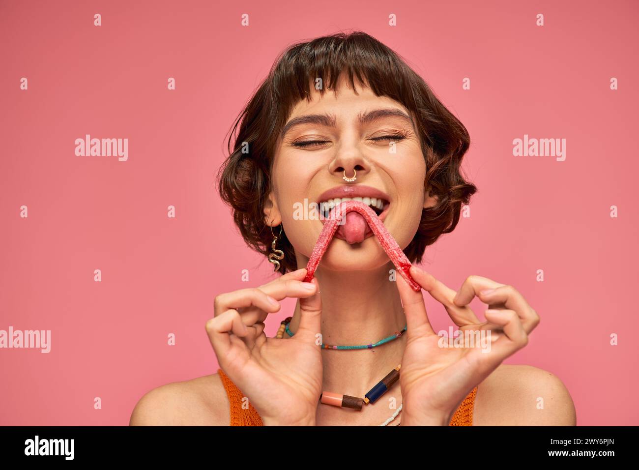 pleased and young woman with nose piercing licking sweet and sour candy strip on pink background Stock Photo