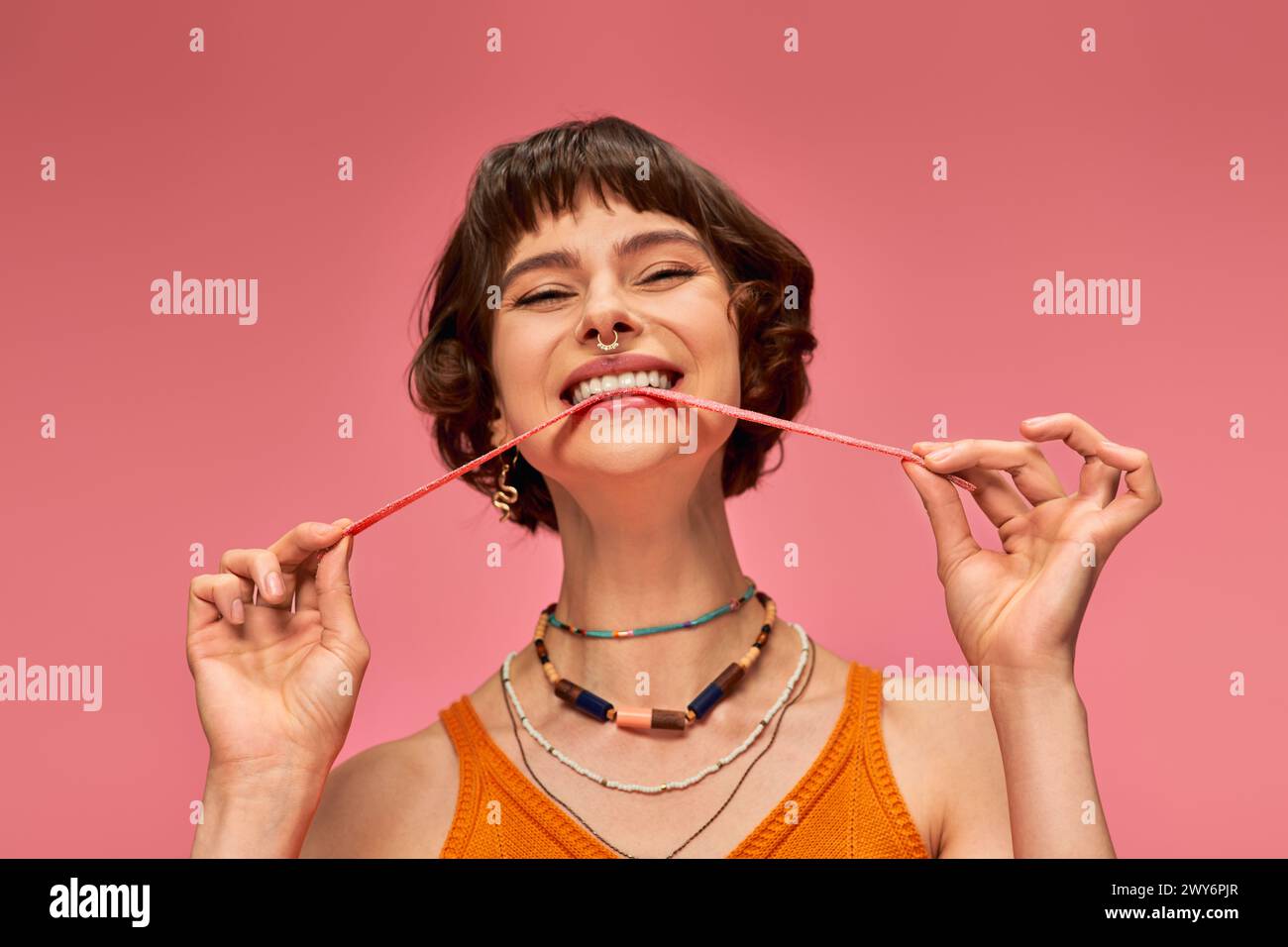 delighted and young woman with nose piercing biting sweet and sour candy strip on pink background Stock Photo