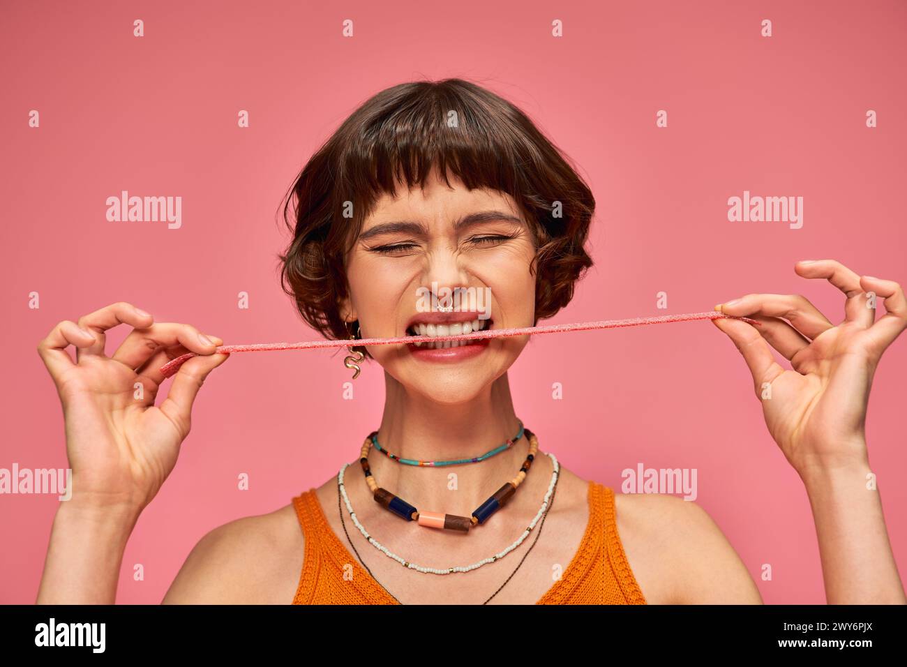 expressive and young woman with nose piercing biting sweet and sour candy strip on pink background Stock Photo