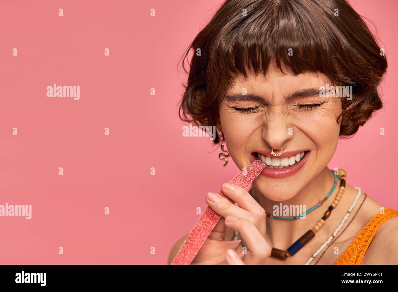expressive woman with nose and white teeth biting sweet and sour candy strip on pink Stock Photo