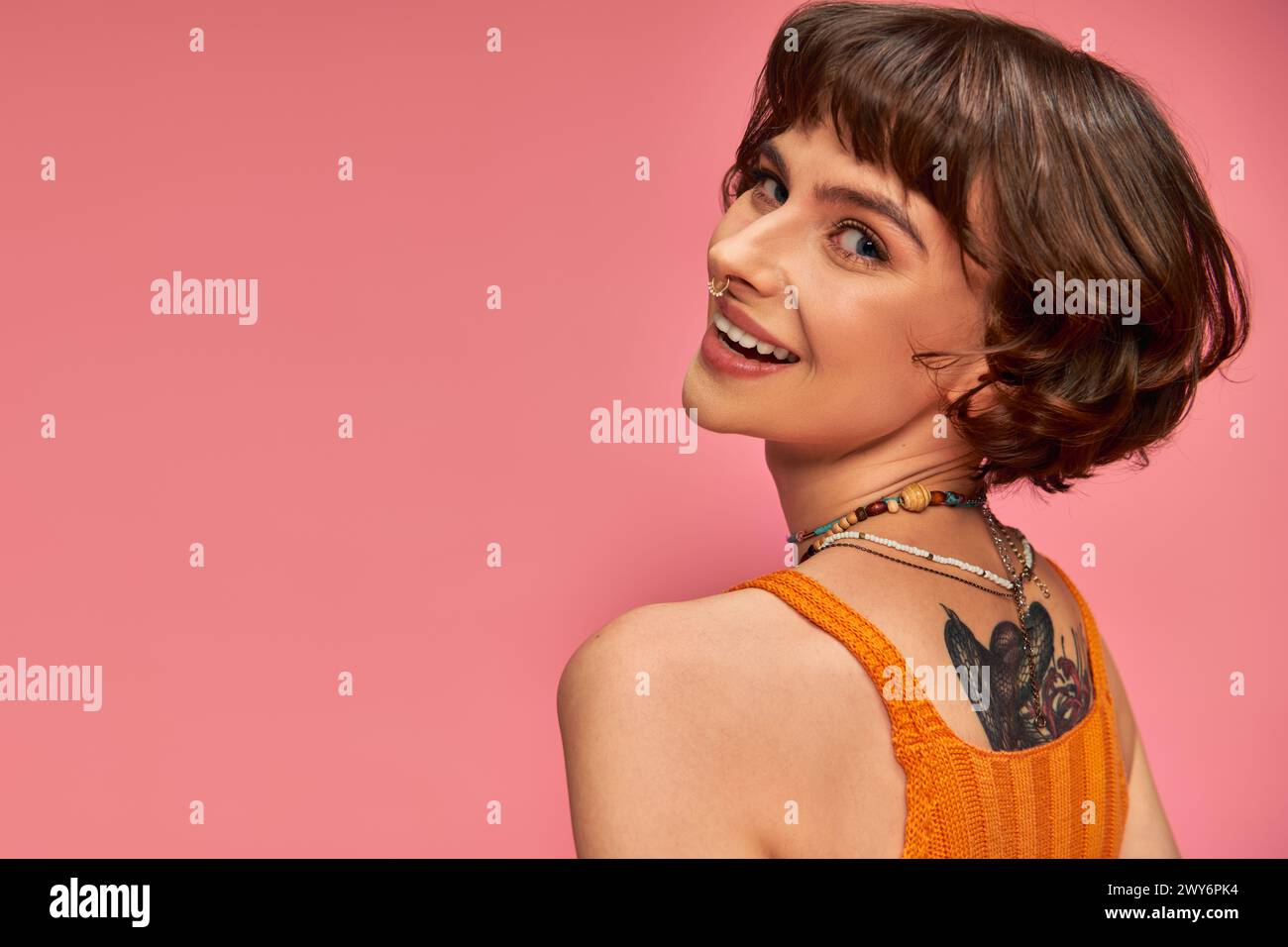 happy tattooed woman in 20s with nose piercing and brunette short hair smiling on pink background Stock Photo