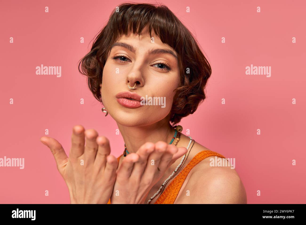 flirty young woman with nose piercing and short brunette hair sending air kiss on pink background Stock Photo