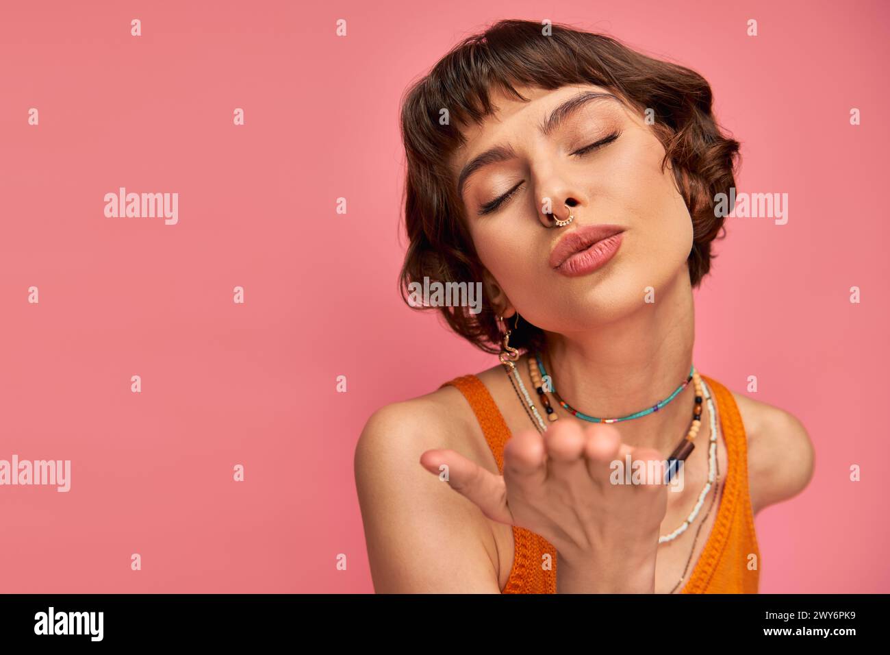 flirty young woman with nose piercing and short brunette hair showing kiss on pink background Stock Photo