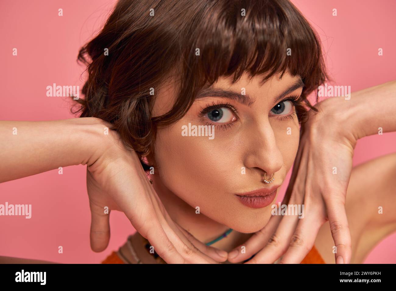 flirty young woman with nose piercing and short brunette hair pouting lips on pink background Stock Photo