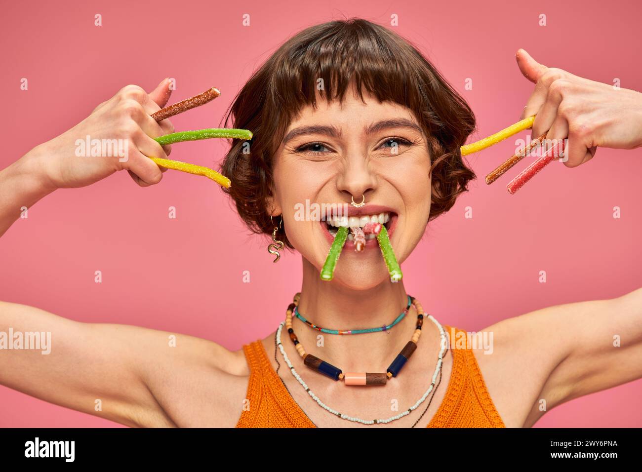 happy young woman in her 20s with sweet and sour candies in hands and teeth on pink background