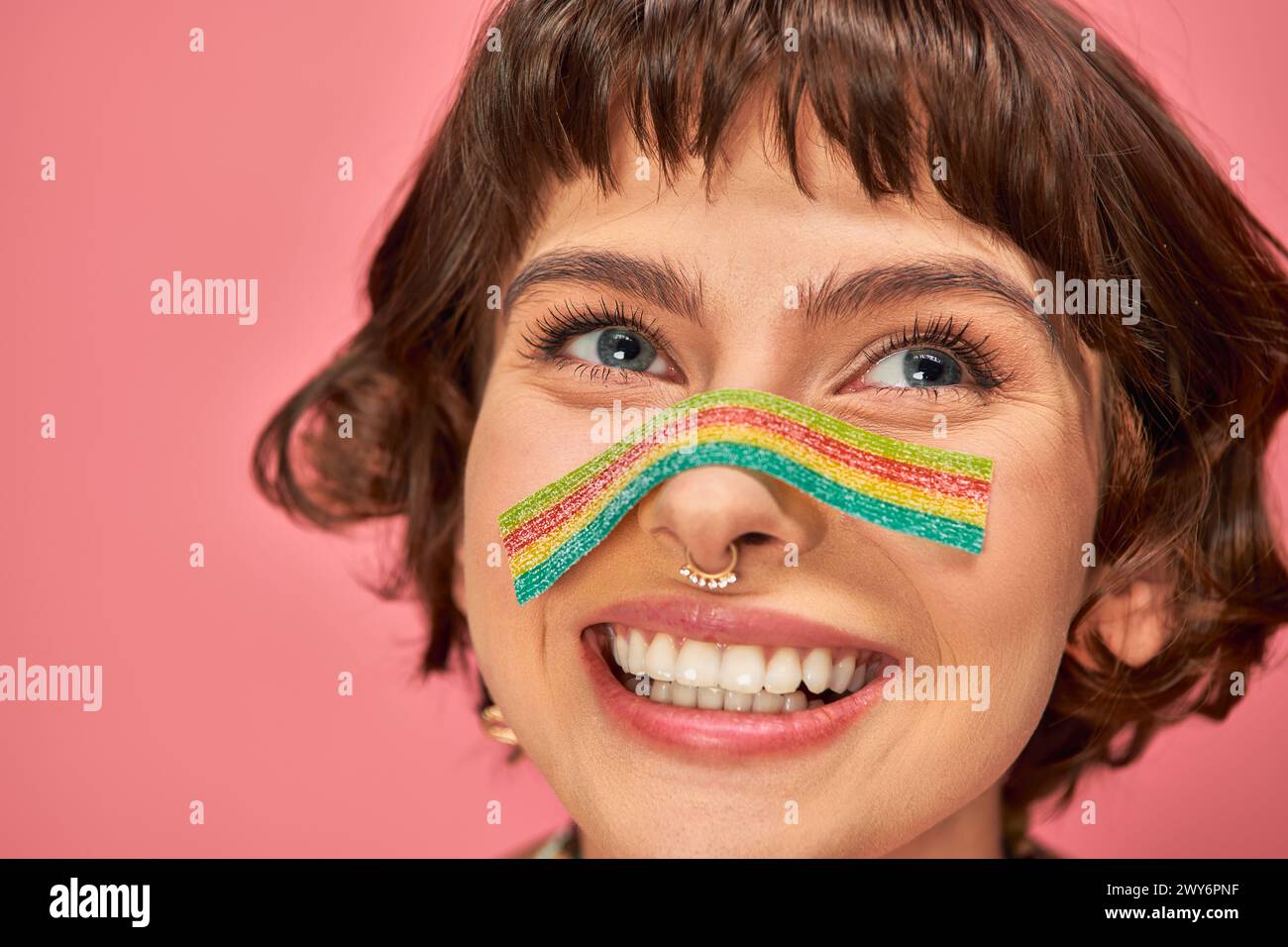 happy woman in her 20s with sweet and sour candy strip aon her nose on pink background Stock Photo