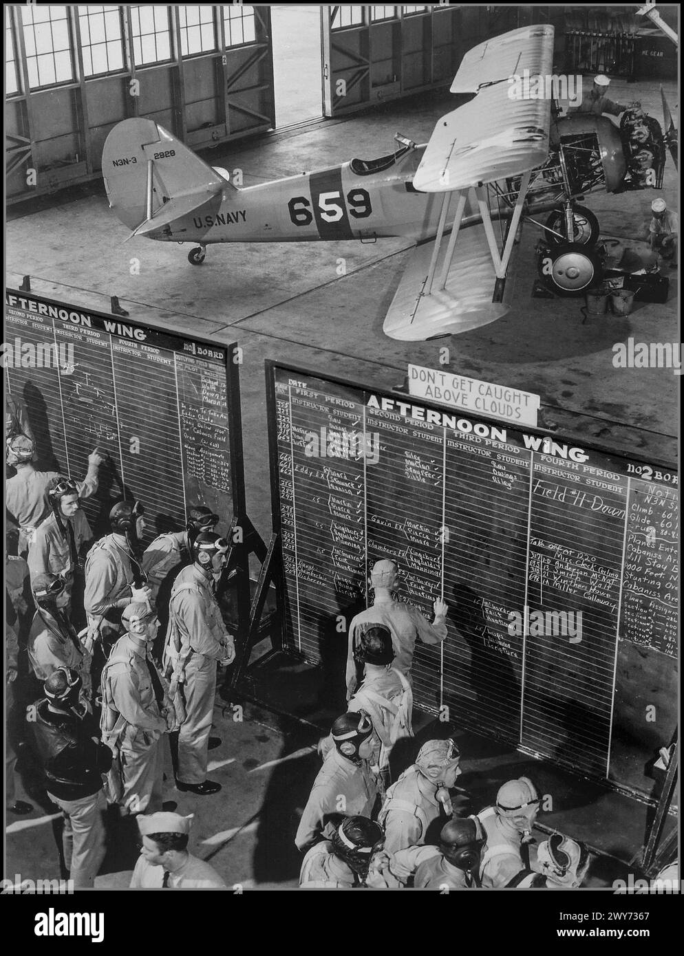 WW2 1942 USA Cadets of the American aviation school at the Naval Air Station Corpus Christi at the training flight schedule. In the background of the photo is a Naval Aircraft Factory N3N-3 training aircraft (serial number 2928, tail number 659). Schedule training blackboards, with cautionary message 'DONT GET CAUGHT ABOVE THE CLOUDS'  Corpus Christi USA Stock Photo