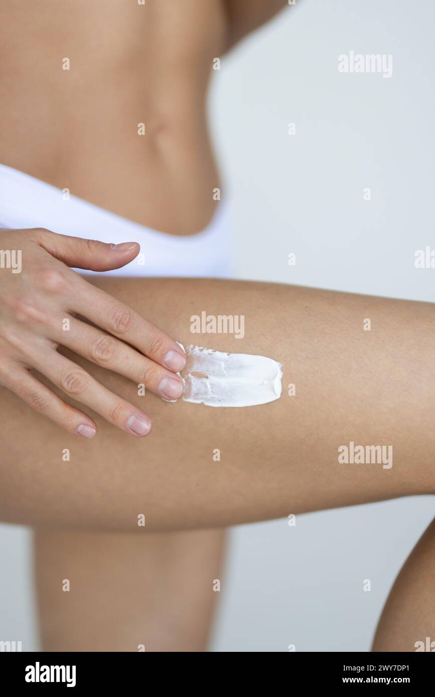 Brunette woman in white underwear applying cream on leg on grey Stock Photo