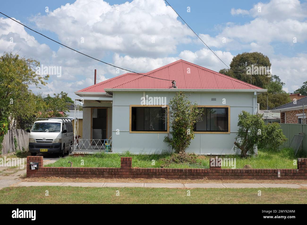 A lowset fibro house with a red corrugated iron roof, trees, shrubs and clouds in the Western Sydney suburb of Canley Vale Stock Photo