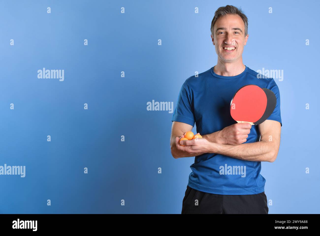 Smiling man in sportswear showing ping pong paddles and balls in hands on blue isolated background. Stock Photo
