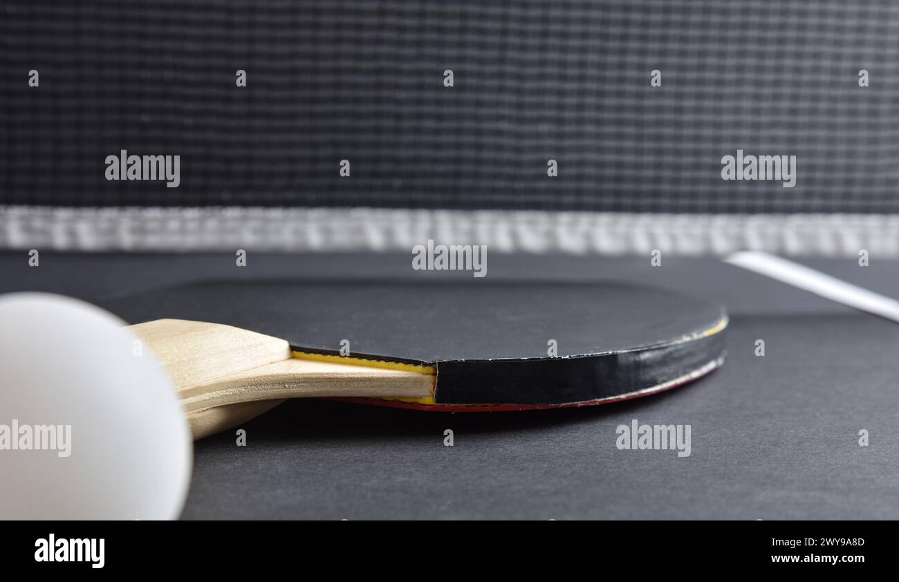 Detail of ping pong paddle and ball with black rubber on black playing table and net in the background. Front view. Stock Photo