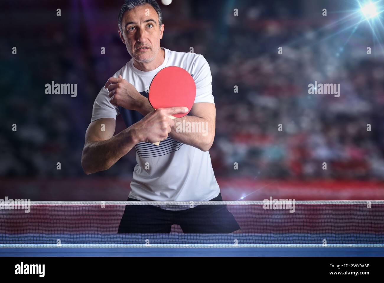 Attentive ping-pong player receiving a white ball behind a blue game table in a exhibition with stands in the background. Stock Photo