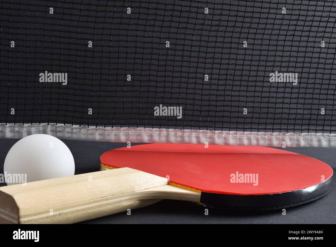 Ping pong set with paddle with red rubber and white ball on a black game table with net in the background. Front view. Stock Photo