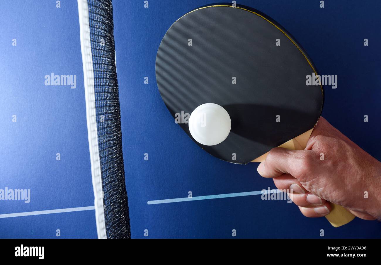 Detail of hand with table tennis paddle hitting a white ball next to the net on a blue game table. Top view. Stock Photo