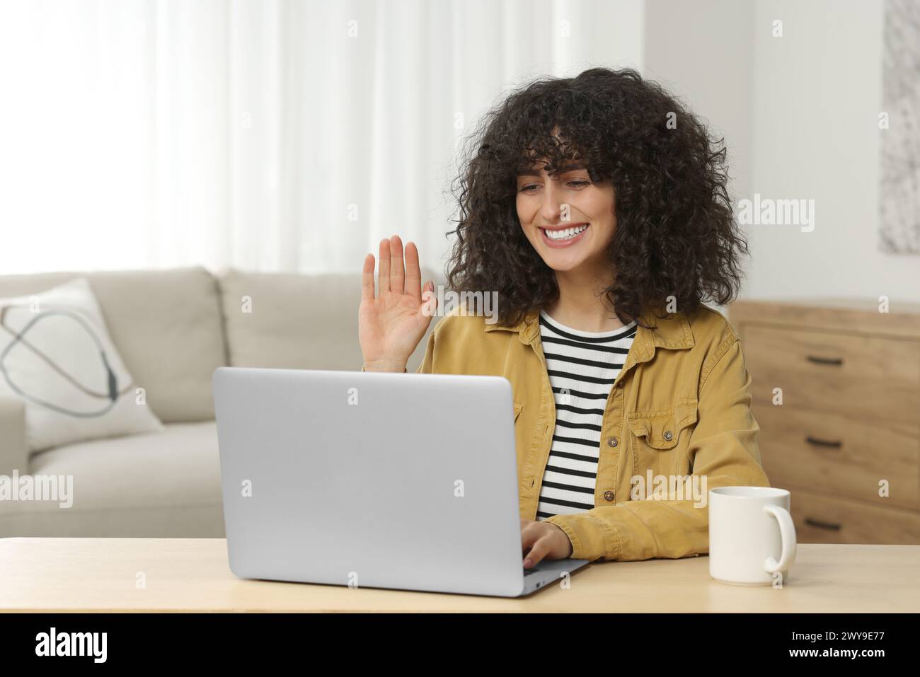 Happy woman waving hello during video call at table in room Stock Photo