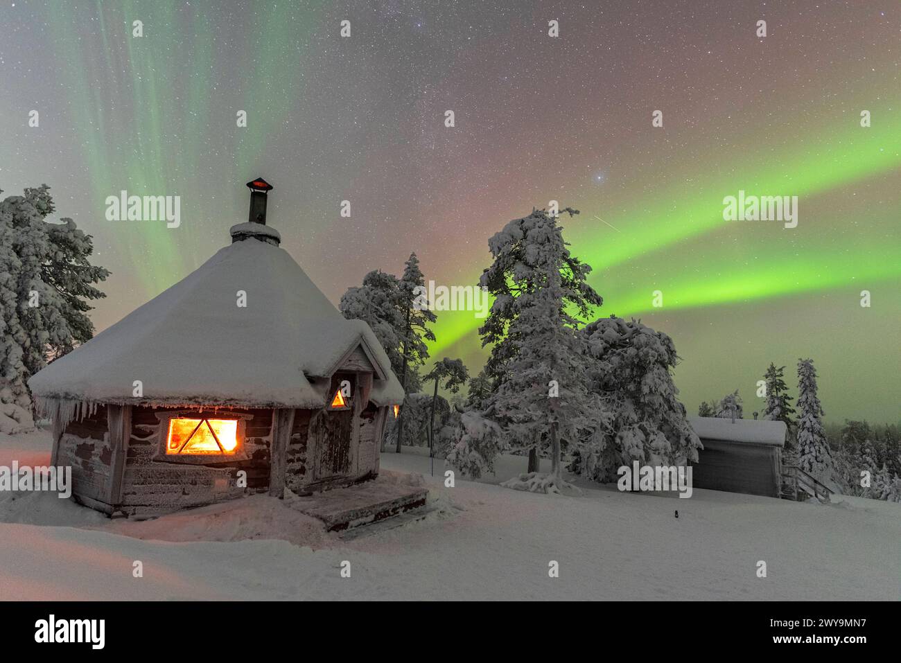 Shades of green color the Northern Lights Aurora Borealis above a snowy landscape with a typical hut lit by a fire inside in the foreground, Finnish L Stock Photo