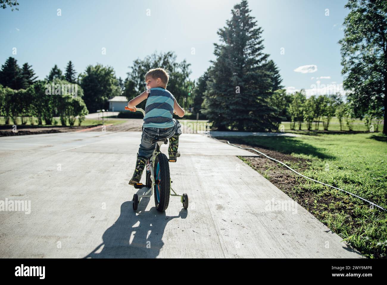 Rear view of boy riding bike with training wheels in drive in summer