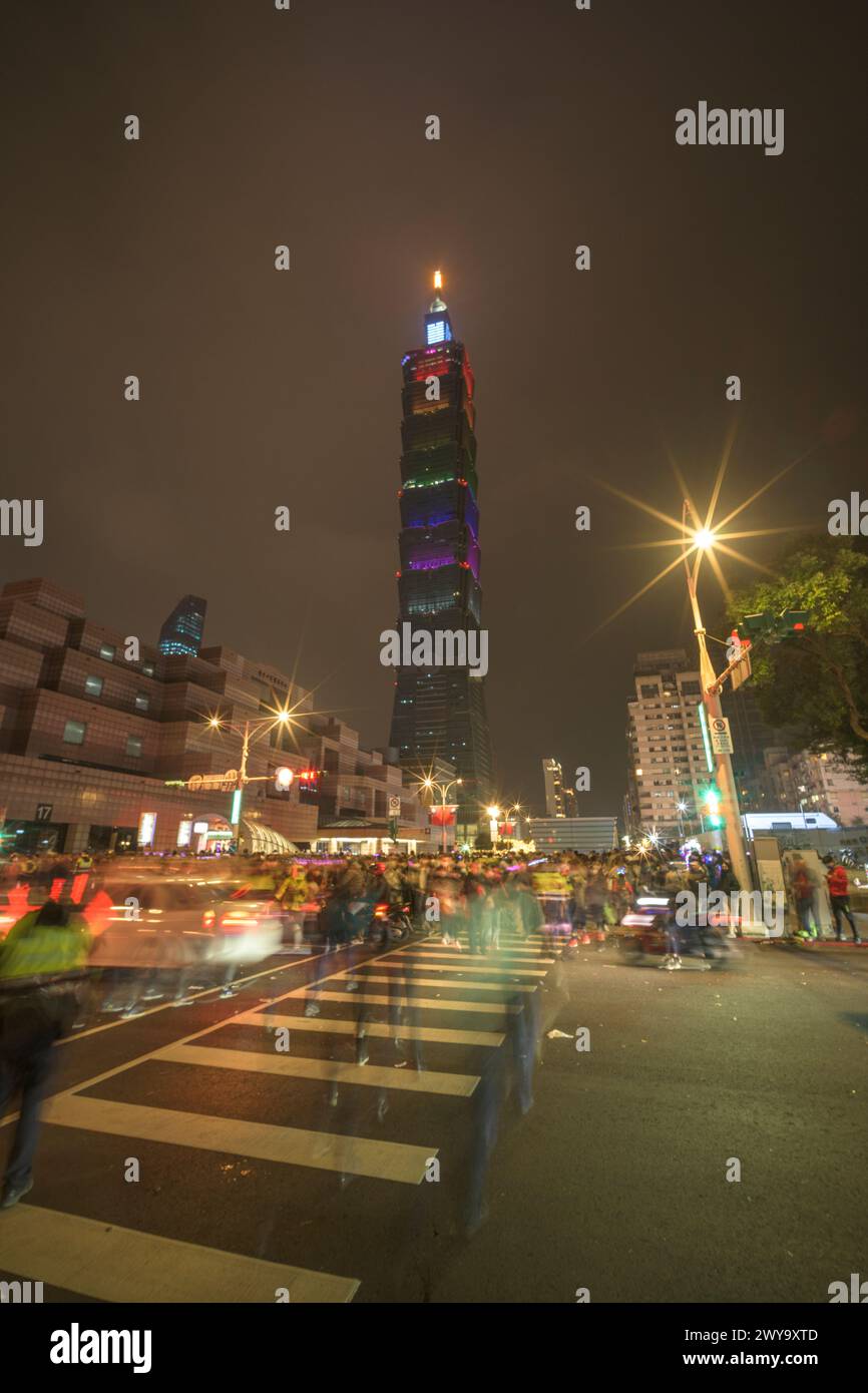 Bustling street scene with moving traffic and pedestrians leads to the illuminated Taipei 101 tower against the dark sky after new year's eve fireworks show Stock Photo