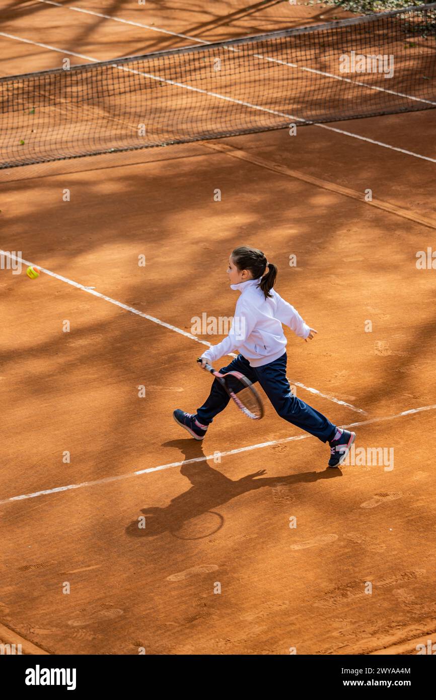 A little girl plays tennis on a red clay court. She runs and trains to learn the beautiful sport of tennis. Competitiveness, athlete, sporty child. Stock Photo