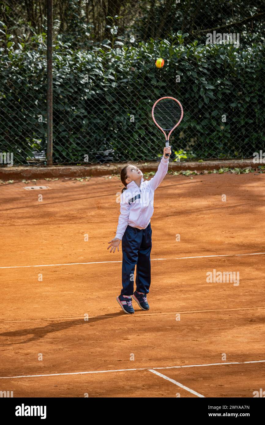 A little girl plays tennis on a red clay court. She runs and trains to learn the beautiful sport of tennis. Competitiveness, athlete, sporty child. Stock Photo
