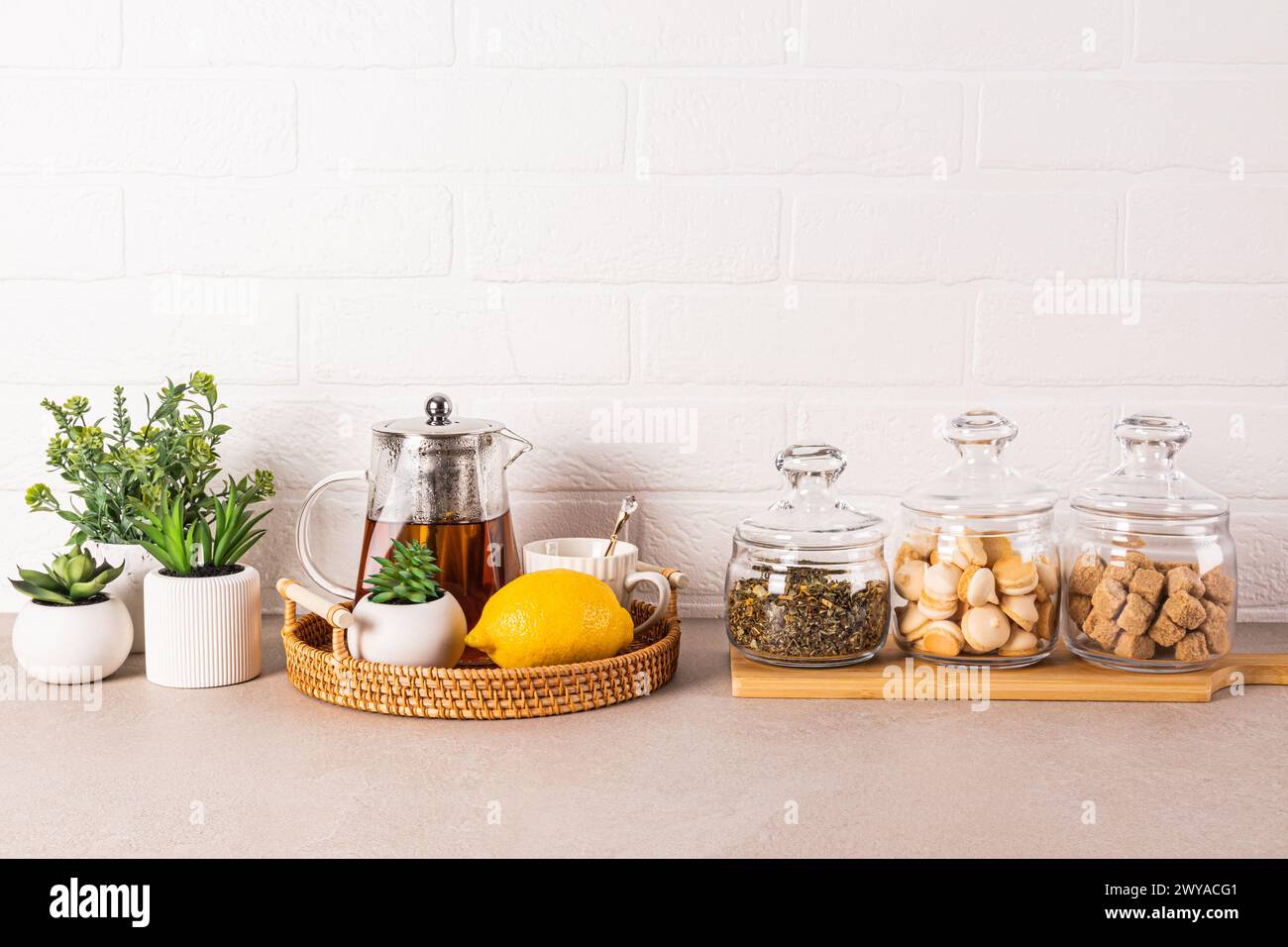 Stylish kitchen background with glass tea pot, lemon, cup, tea jars, sugar, meringue pastry. Green plants in pots. Front view Stock Photo