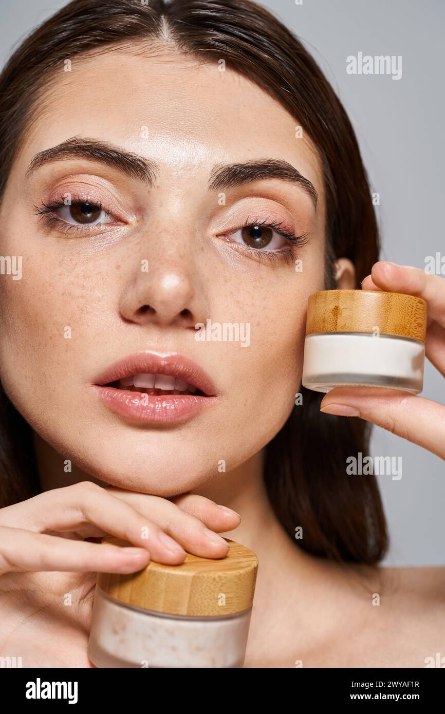 A young Caucasian woman with brunette hair applies cream from a jar to her face, enhancing her natural beauty. Stock Photo