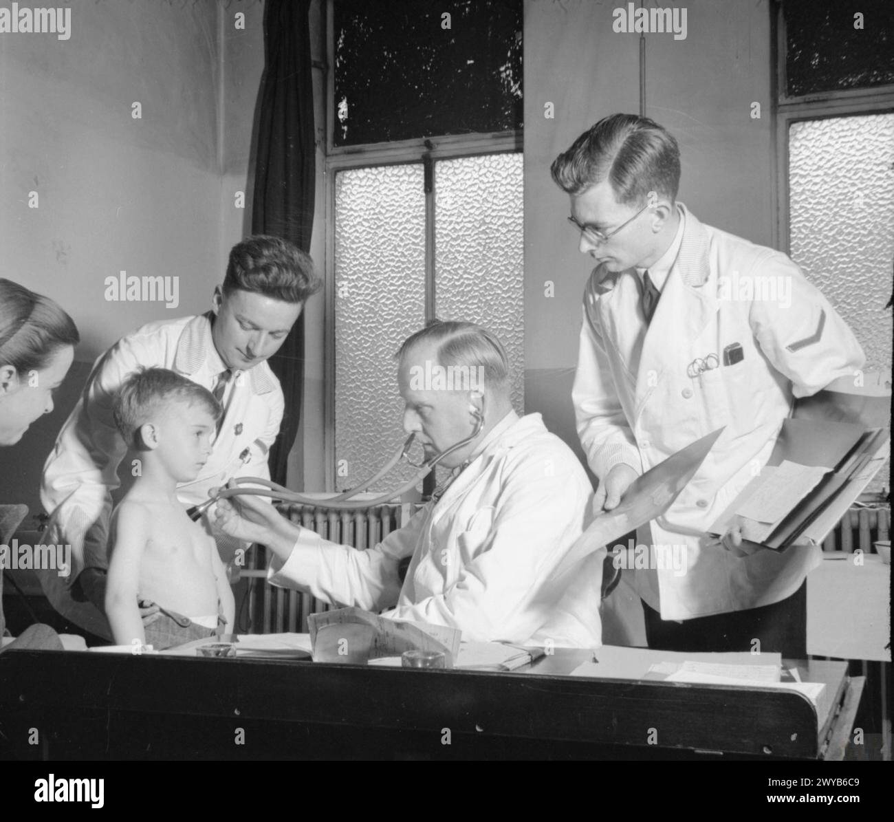 MALE NURSES: LIFE AT HACKNEY HOSPITAL, LONDON, 1943 - Male nurses assist during a chest examination as part of their training at Hackney Hospital in London. One nurse holds the young boy in the correct position as the doctor listens to his chest with his stethoscope. A second nurse shows the doctor an x-ray of the patient's chest. The boy's mother is just visible on the left of the photograph. , Stock Photo