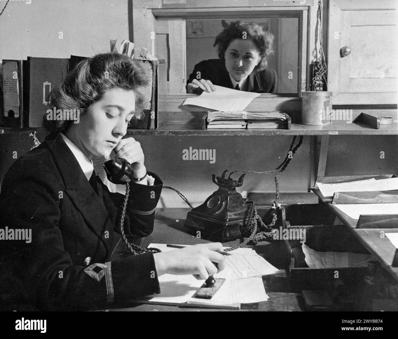 WOMEN'S ROYAL NAVAL SERVICE: WRENS WITH THE FLEET MAIL, ENGLAND, UK, NOVEMBER 1944 - The WRNS Duty Cipher Officer (speaking on the telephone) is hard at work in the Signal Distribution Office of the Central Communications Branch. She is distributing signals for the base. A WRNS Cipher Officer is handing the DCO a deciphered signal through the hatch. , Stock Photo