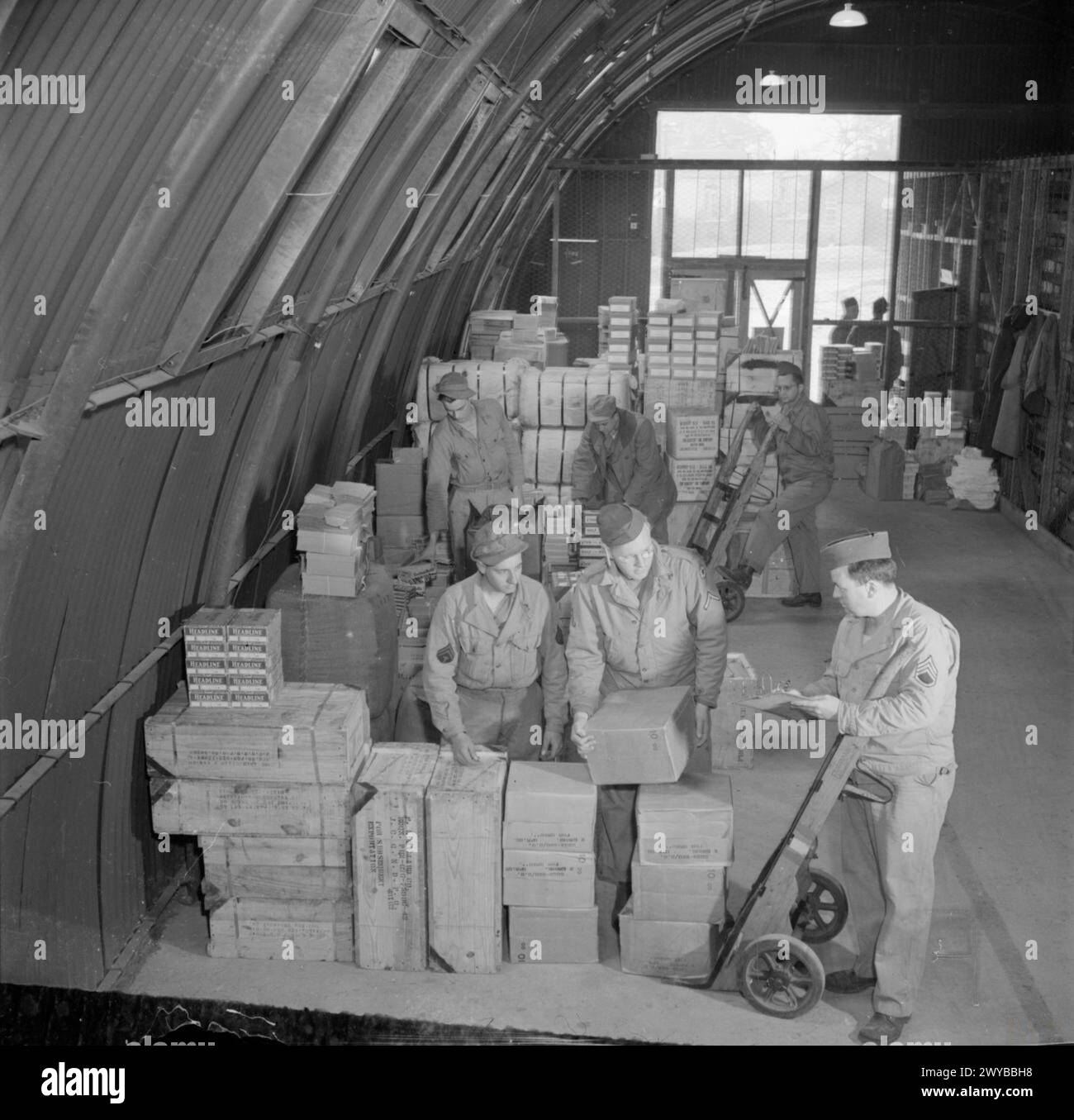BRITAIN SUPPLIES US ARMY STORE: AMERICANS IN BRITAIN, 1943 - GIs at work at an American Army store, somewhere in Britain. This particular area of the store supplies goods for Post Exchanges (also known as PXs), including candles, chocolate, shaving cream, toothpaste, batteries, ink, writing paper, shoe polish and playing cards. Making up a load for dispatch in the foreground are (left to right): T/5 Joe Brethauer (of Rote 2, Box 245, Fort Lupton, Colorado), Pfc Bernard Hanson (of 148 Magnolia Avenue, Kearny, New Jersey) and Staff Sergeant Robert McKenna of Big Bend, Wisconsin. , Stock Photo