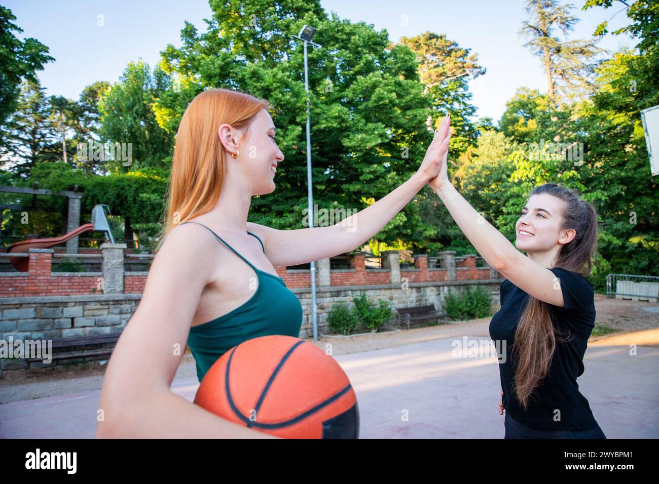 Two women are high fiving each other in a park. One of them is holding a basketball Stock Photo