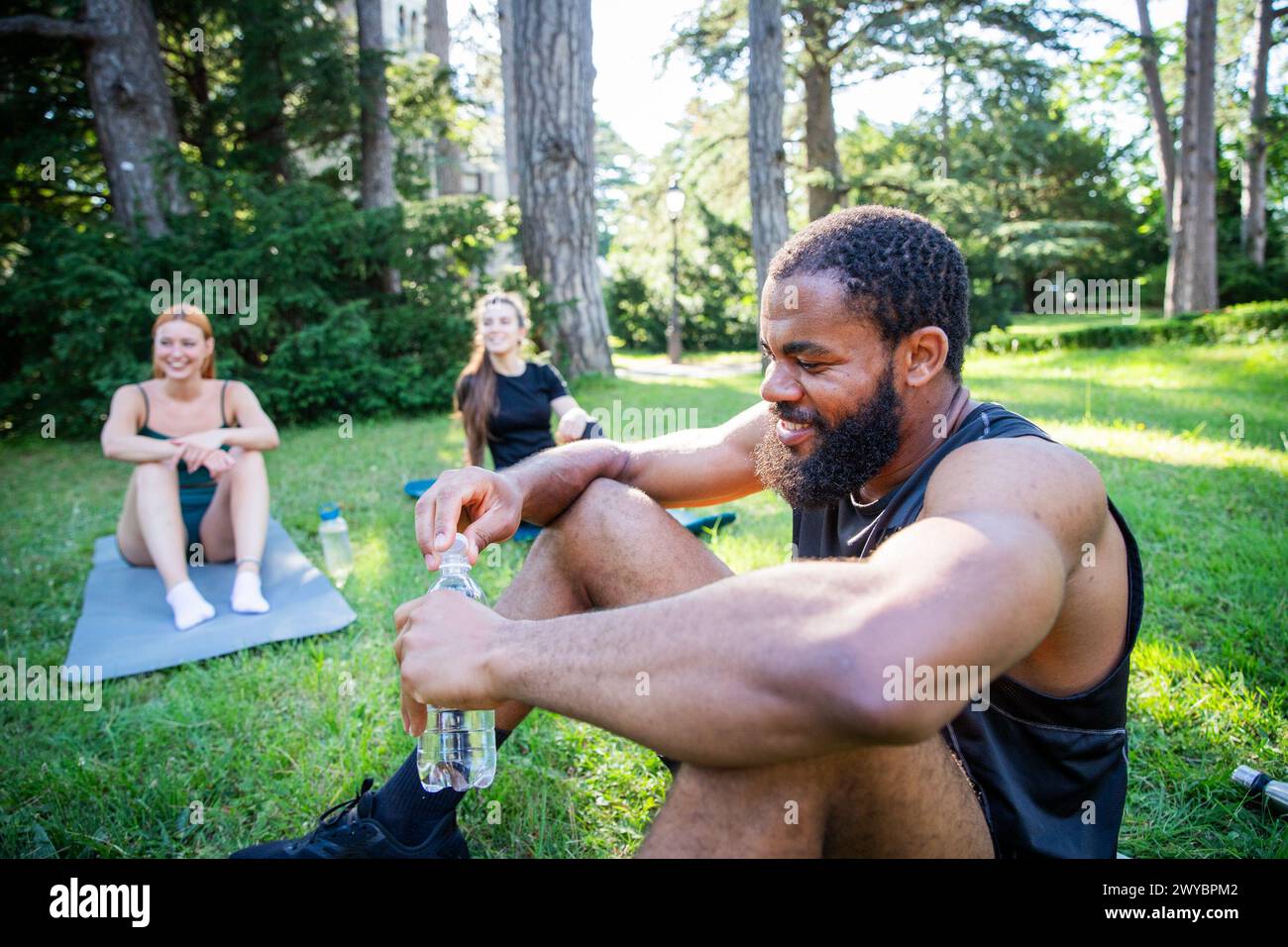 A man with a beard is sitting on a mat in a park is drinking water with two other people. Stock Photo