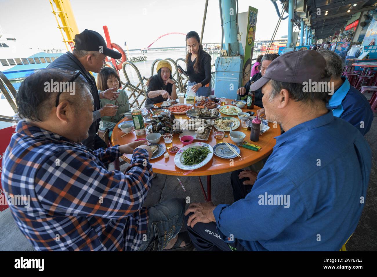 A lively group of friends or family sharing a variety of seafood dishes at an outdoor table by the seaside Stock Photo
