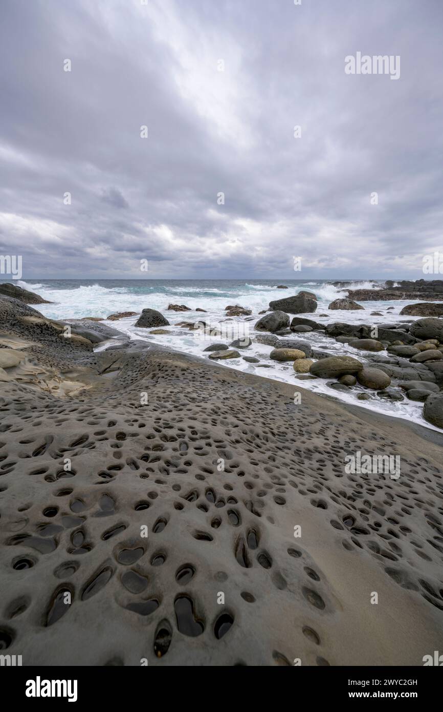 An overcast day on a rocky shore showcasing unique erosion  hole patterns with rough waves in the background Stock Photo