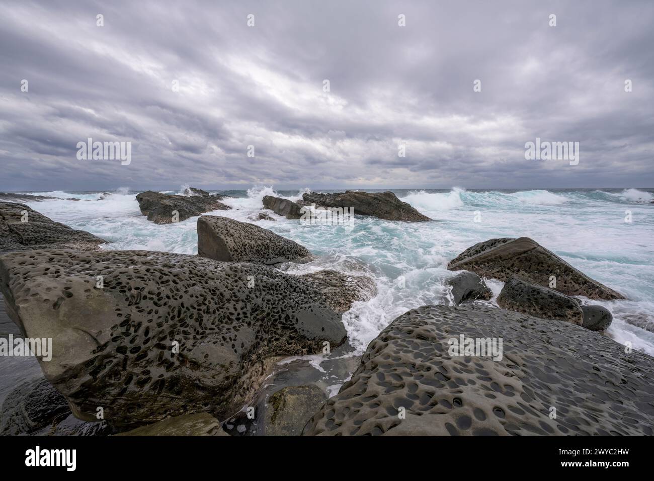 An overcast day on a rocky shore showcasing unique erosion  hole patterns with rough waves in the background Stock Photo